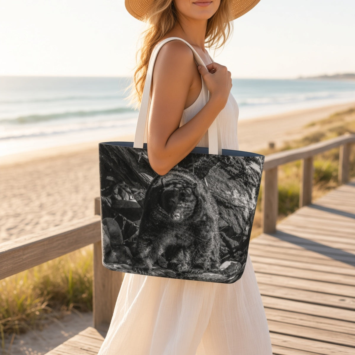 A woman on a wooden boardwalk by the beach carries a large tote bag with a black-and-white animal print. The tote has white shoulder straps and a dark navy border.