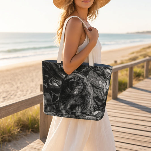 A woman on a wooden boardwalk by the beach carries a large tote bag with a black-and-white animal print. The tote has white shoulder straps and a dark navy border.