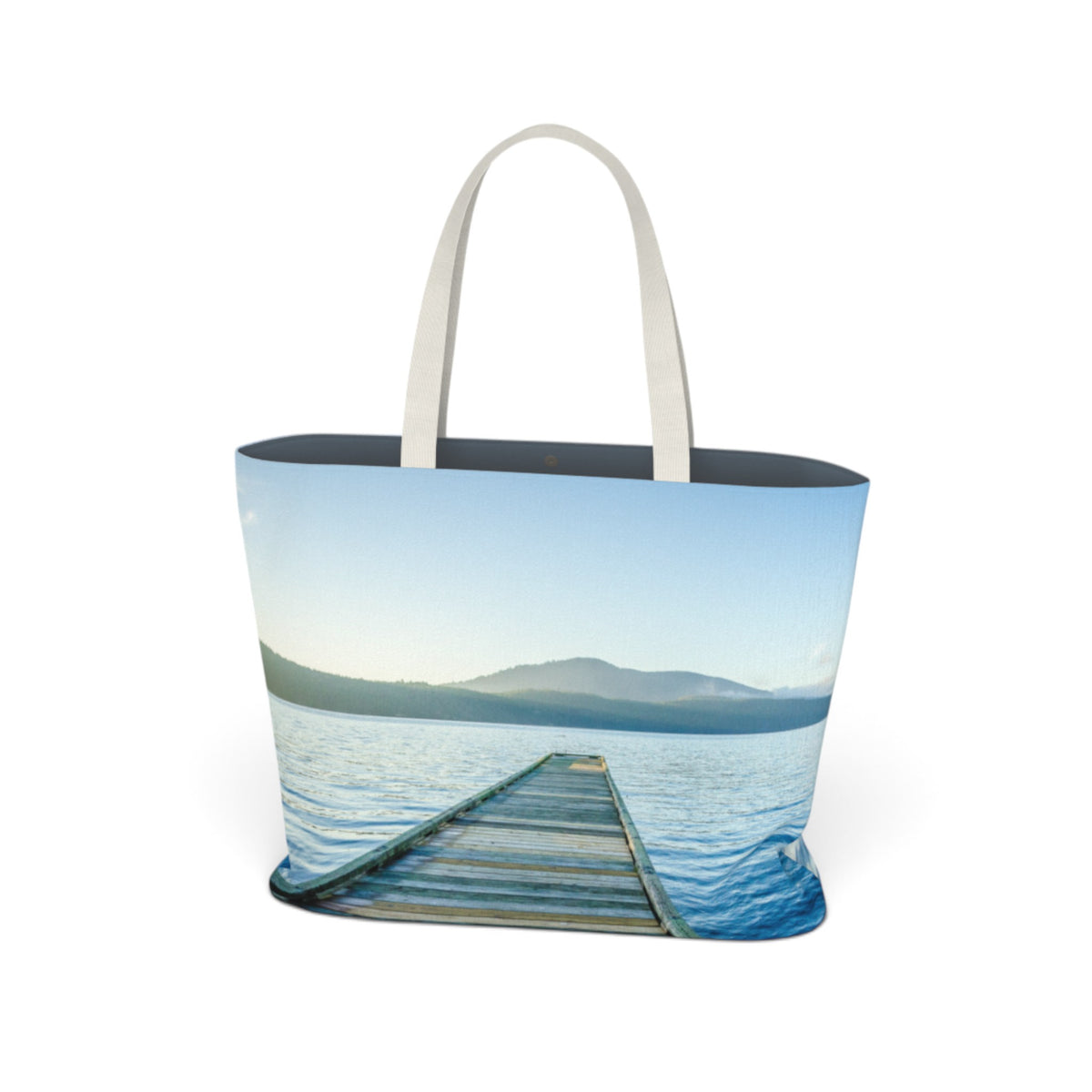 A tote bag featuring a scenic print of a wooden dock extending into calm blue water under a pale sky, with dark blue handles and a dark interior.