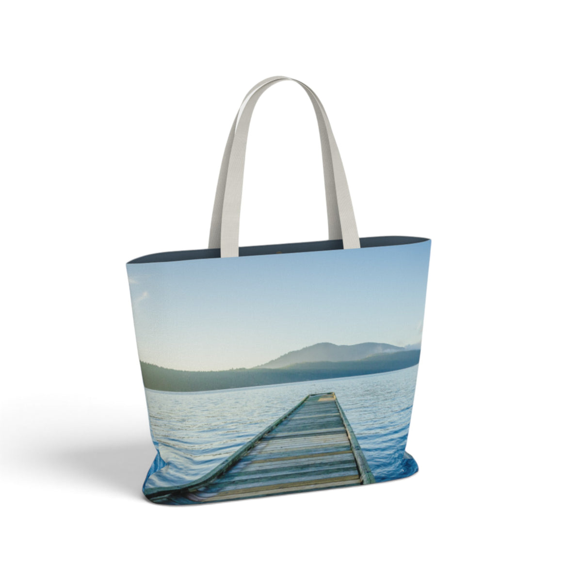 A tote bag featuring a scenic print of a wooden dock extending out over a calm lake with distant mountains under a pale blue sky. The bag has light gray handles and a dark navy blue inner lining, shown on a clean white background.
