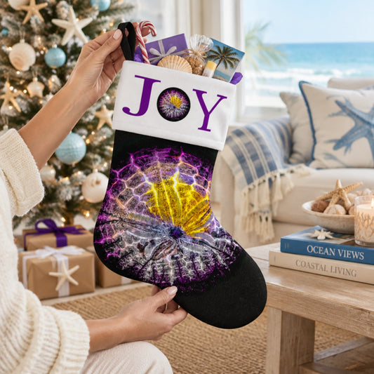 A person holds a black Christmas stocking with purple and white seashell artwork and the word 'JOY' in white on a purple band. The stocking is shown in a cozy living room with a beach-themed couch, wrapped gifts, and a decorated tree in the background.