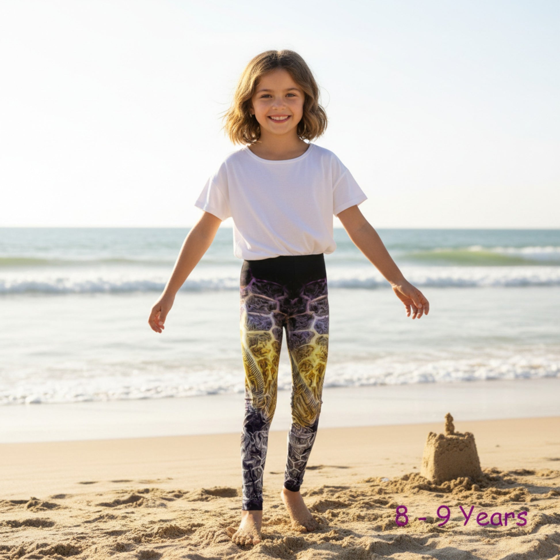 Child wearing colorful leggings and a white shirt on a beach with sandcastle in the background