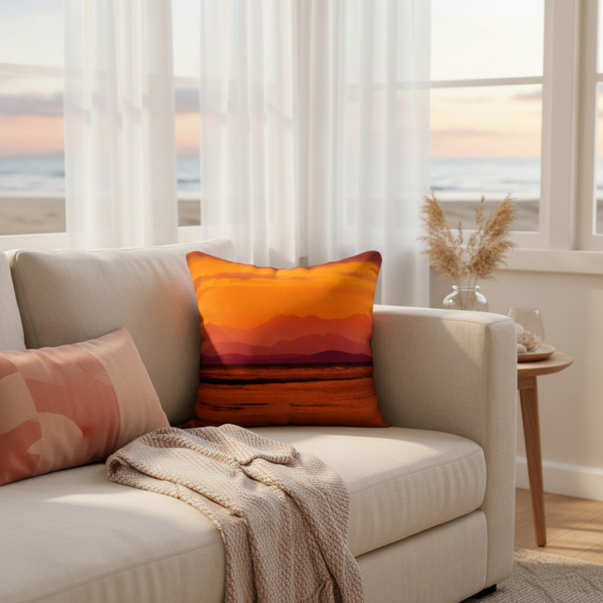 A square throw pillow with a colorful sunset mountain landscape print in orange, yellow, and red, styled on a light beige sofa in a bright living room with an ocean view.
