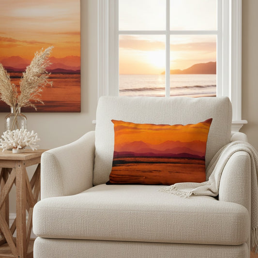 A square throw pillow featuring a vibrant orange and yellow sunset landscape print, styled on a cream-colored armchair in a bright living room setting.