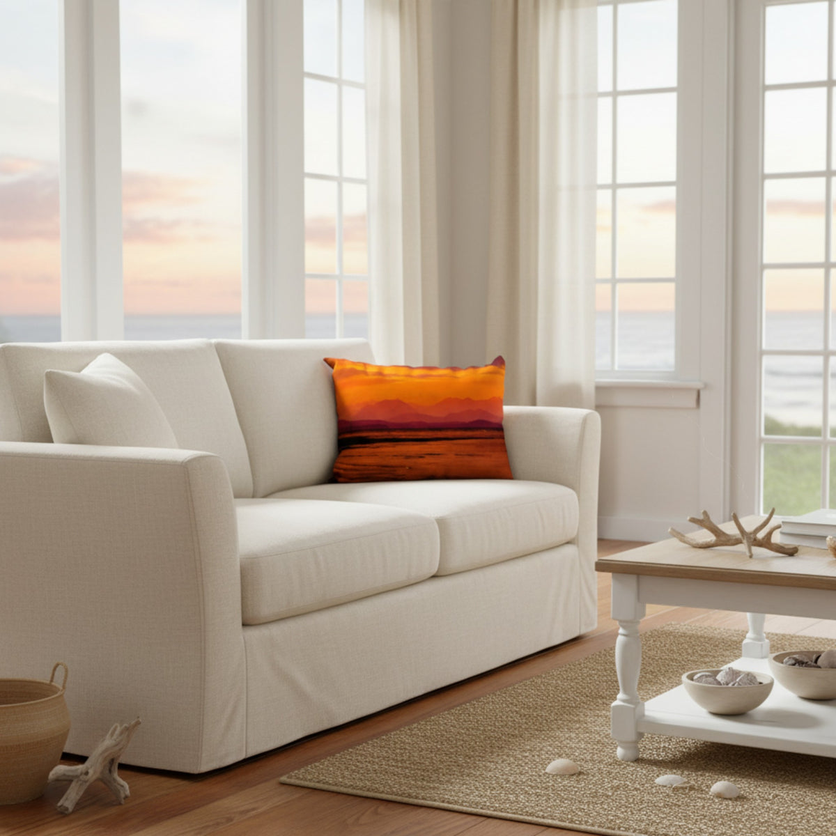 An off-white slipcovered loveseat sofa in a bright, modern living room with large windows overlooking the ocean at sunset. A vibrant orange and red sunset landscape throw pillow is placed on the sofa.