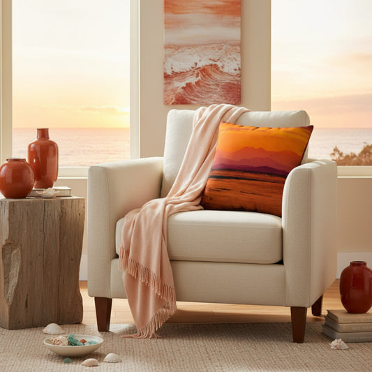 A cream upholstered armchair in a bright coastal living room, styled with a peach throw blanket and a square pillow featuring a sunset landscape. A rustic wood side table with terracotta vases sits beside the chair, and a window shows the ocean at sunset.