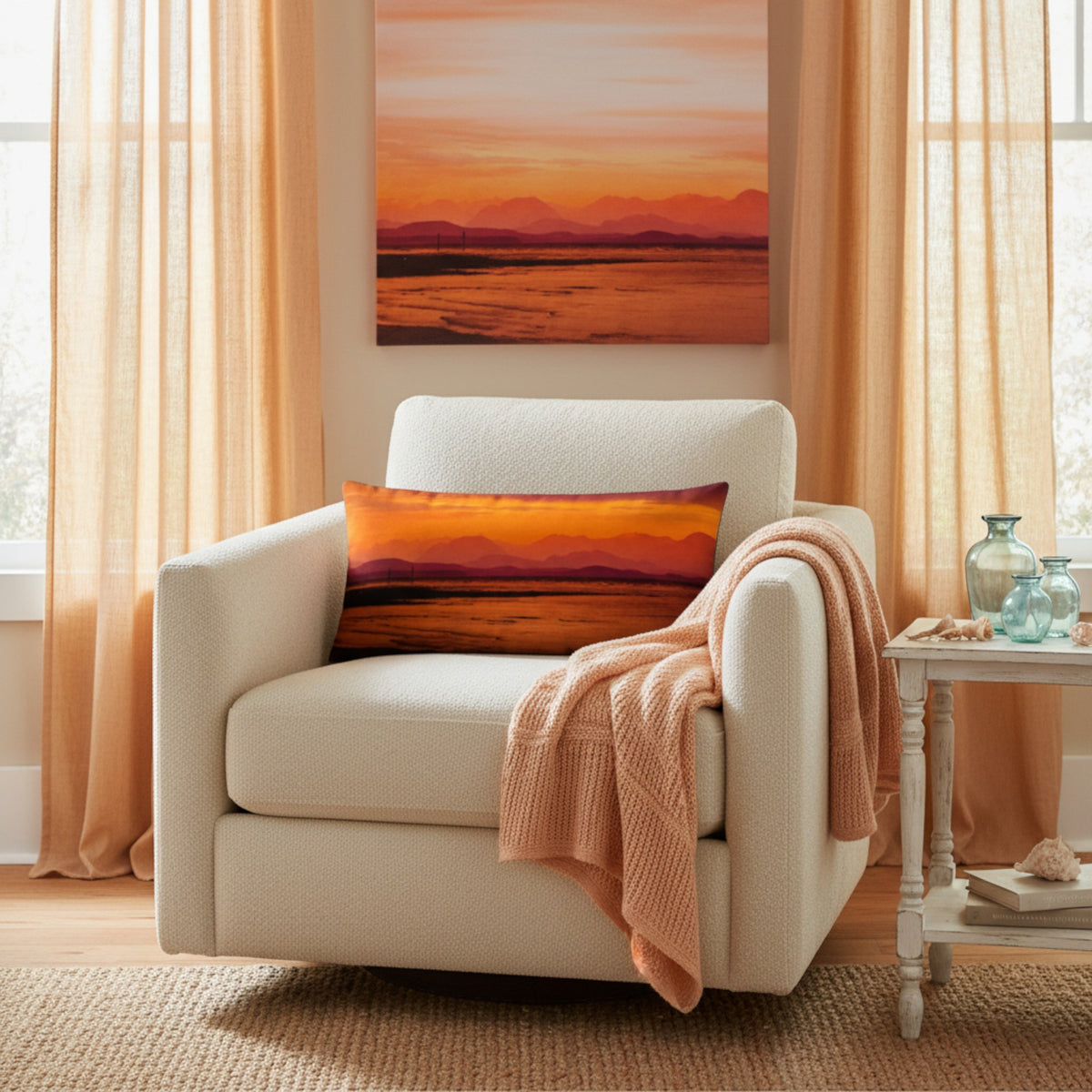A square decorative throw pillow featuring a warm orange and yellow sunset landscape print, styled on a cream armchair in a cozy room setting with a peach knit blanket and a matching landscape canvas on the wall.