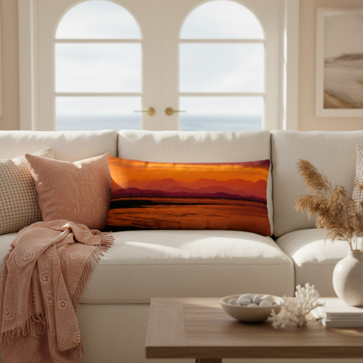 A rectangular throw pillow featuring a vibrant orange and red sunset landscape print, styled on a cream-colored sofa in a bright, modern living room.