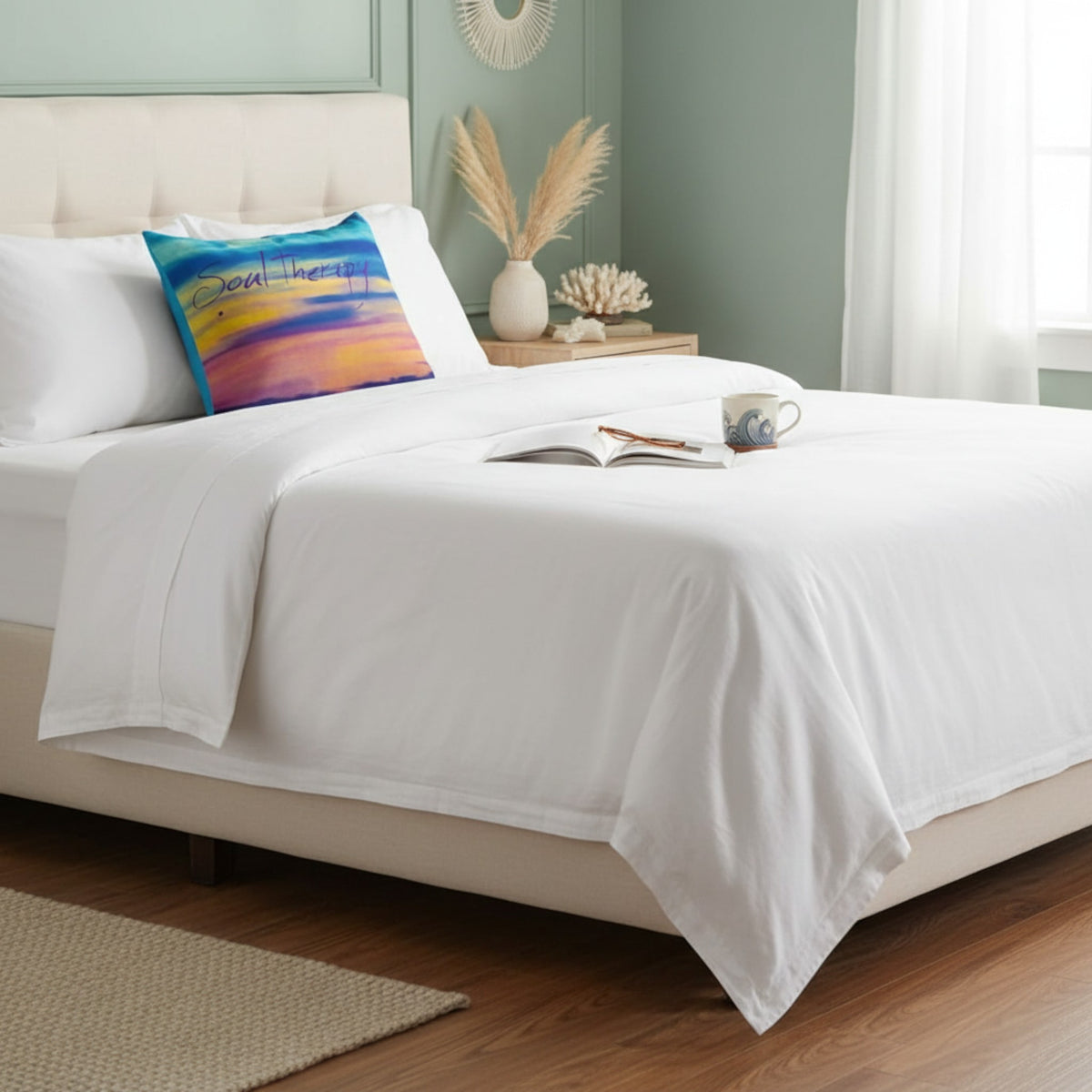 A neatly made bed with crisp white bedding and a colorful abstract throw pillow in a softly styled bedroom with pale green walls, wood flooring, and bedside decor.