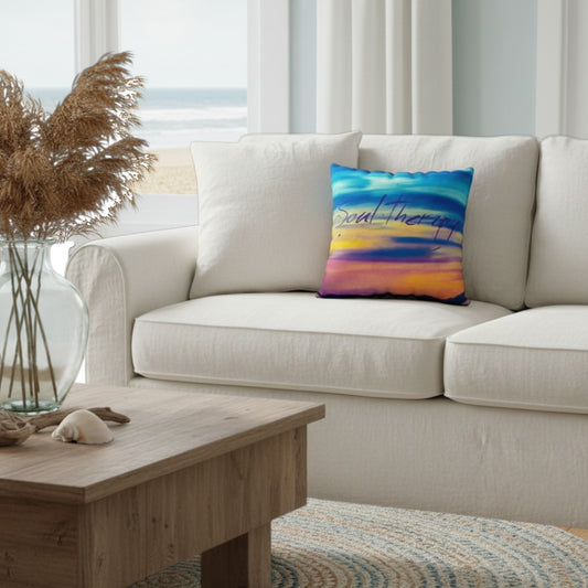 A square throw pillow with a colorful sunset design and the words 'Soal Therapy' printed on it, styled on a cream-colored sofa in a bright, modern living room.