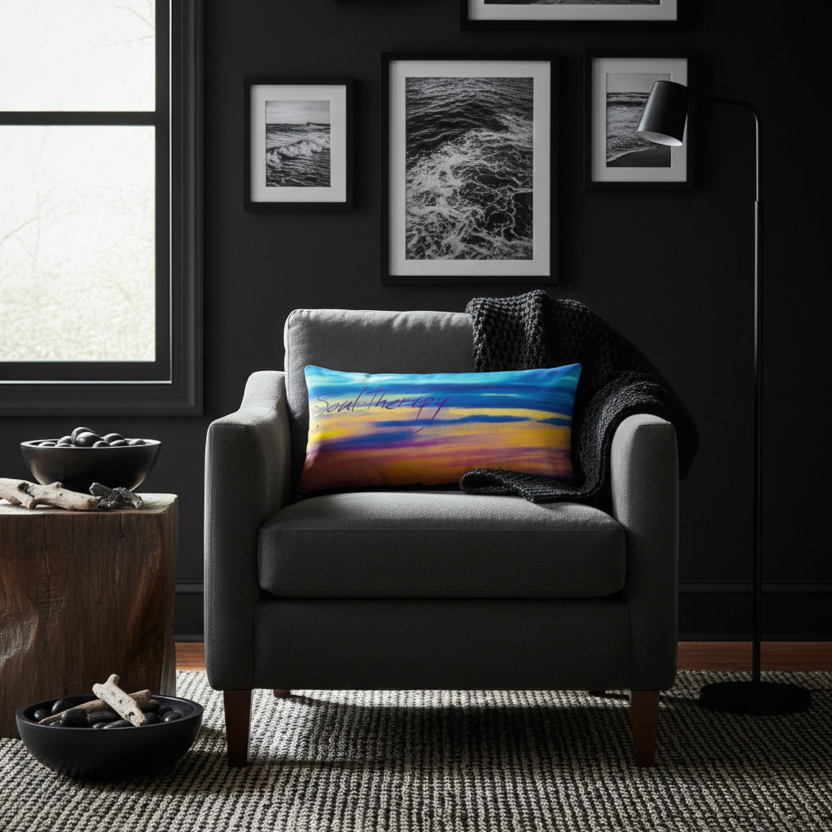 A square throw pillow with a colorful sunset landscape print is styled on a gray armchair in a dark, modern living room setting.