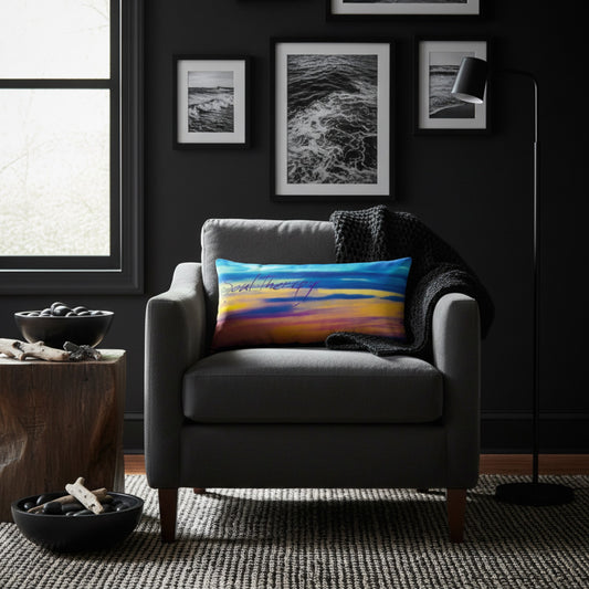 A square throw pillow with a colorful sunset landscape print is styled on a gray armchair in a dark, modern living room setting.