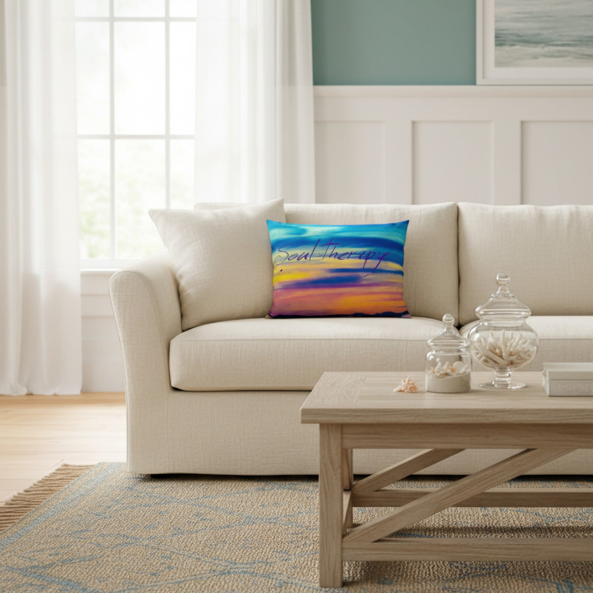 A square throw pillow with a colorful abstract sunset design in blue, orange, yellow, and purple, styled on a cream-colored sofa in a bright, modern living room.