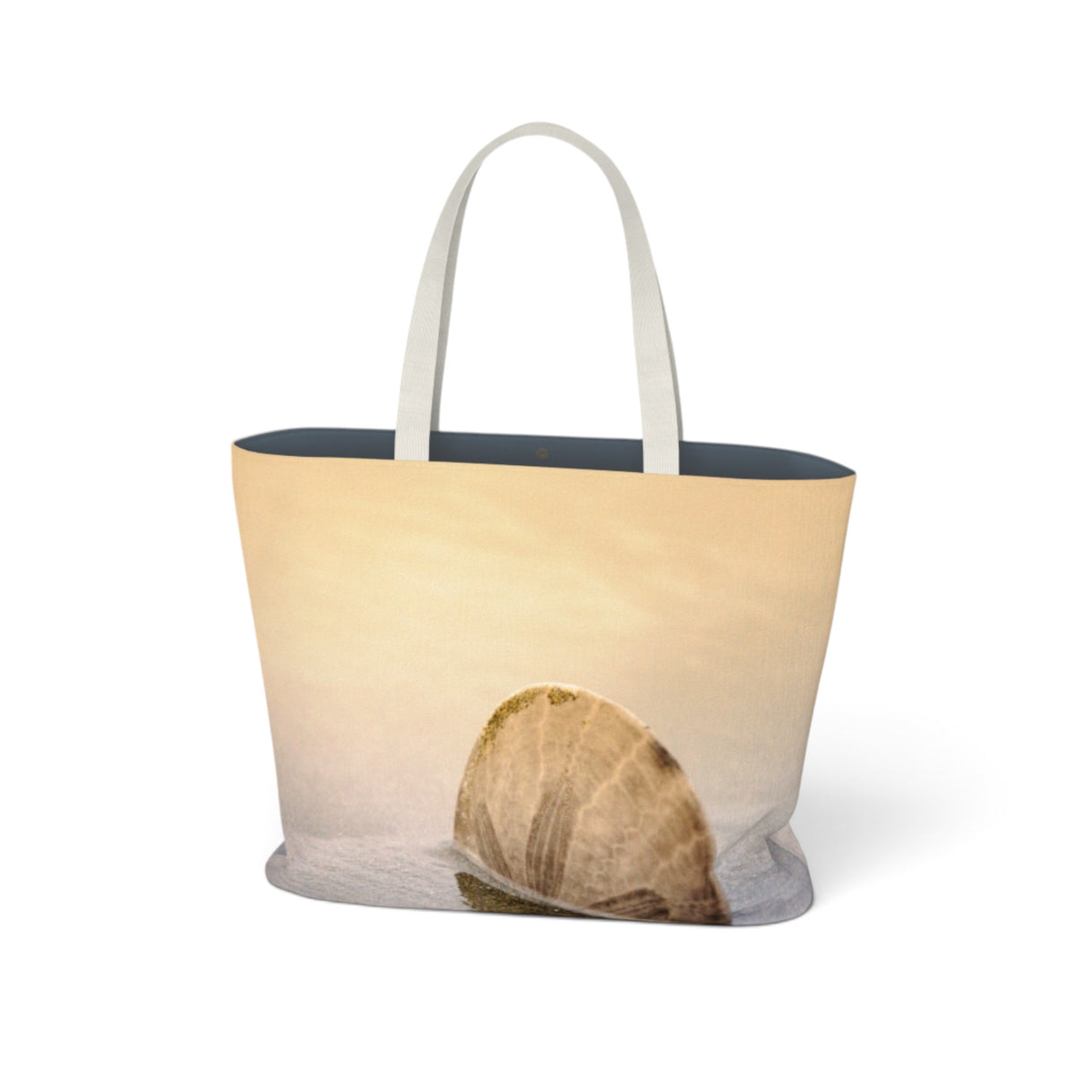 A tote bag featuring a photographic print of a seashell on a sandy beach at sunset, with white shoulder straps and a dark navy blue interior lining, shown against a plain white background.