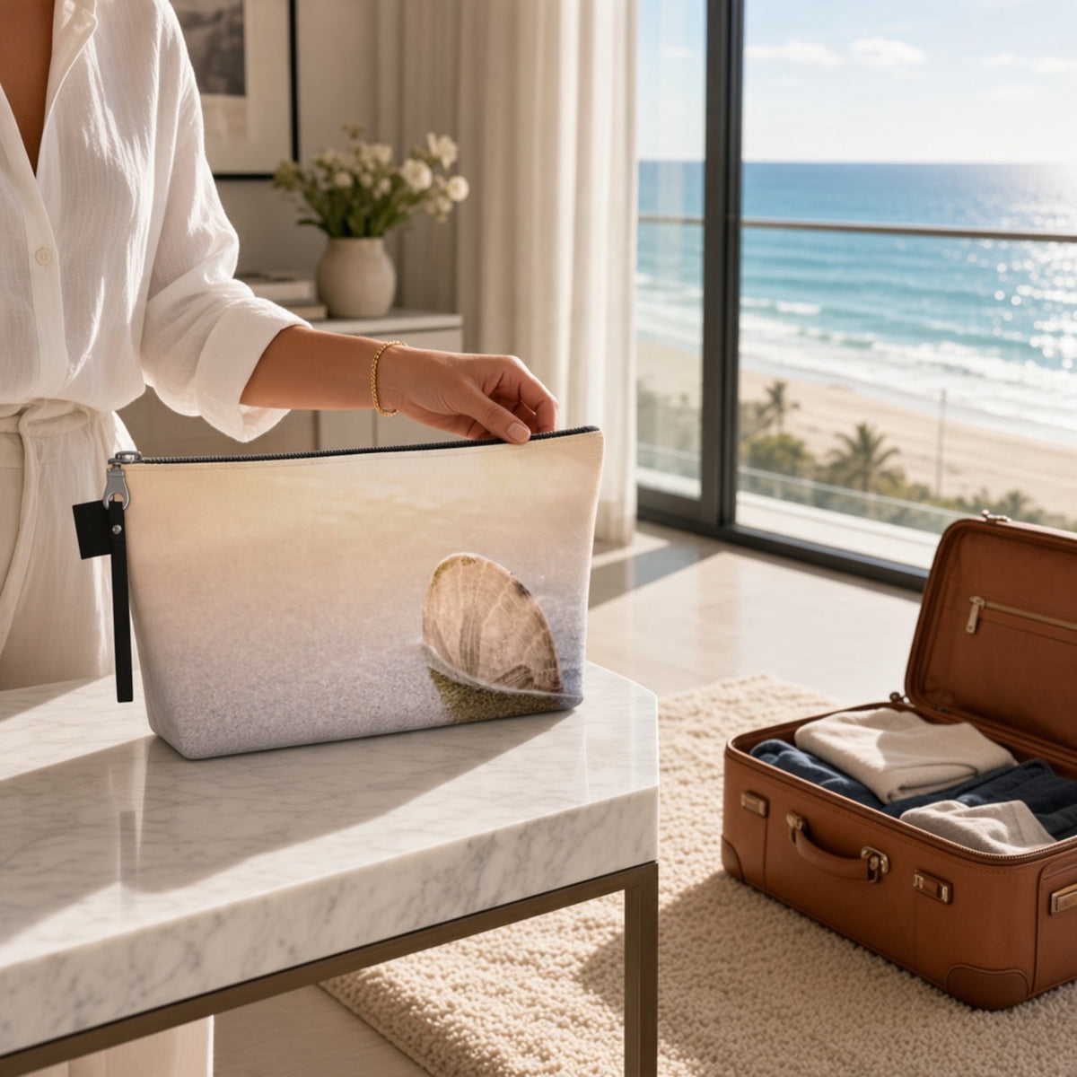 A woman in a white robe places a beach-themed zippered pouch on a marble table next to an open brown suitcase with folded clothes, set against a bright window view of the ocean and beach.