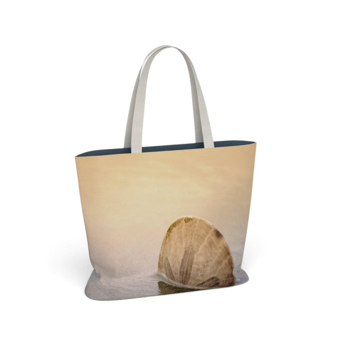 A tote bag featuring a photographic print of a seashell on a sandy beach, with light gray handles and a dark navy blue interior lining, shown against a plain white background.