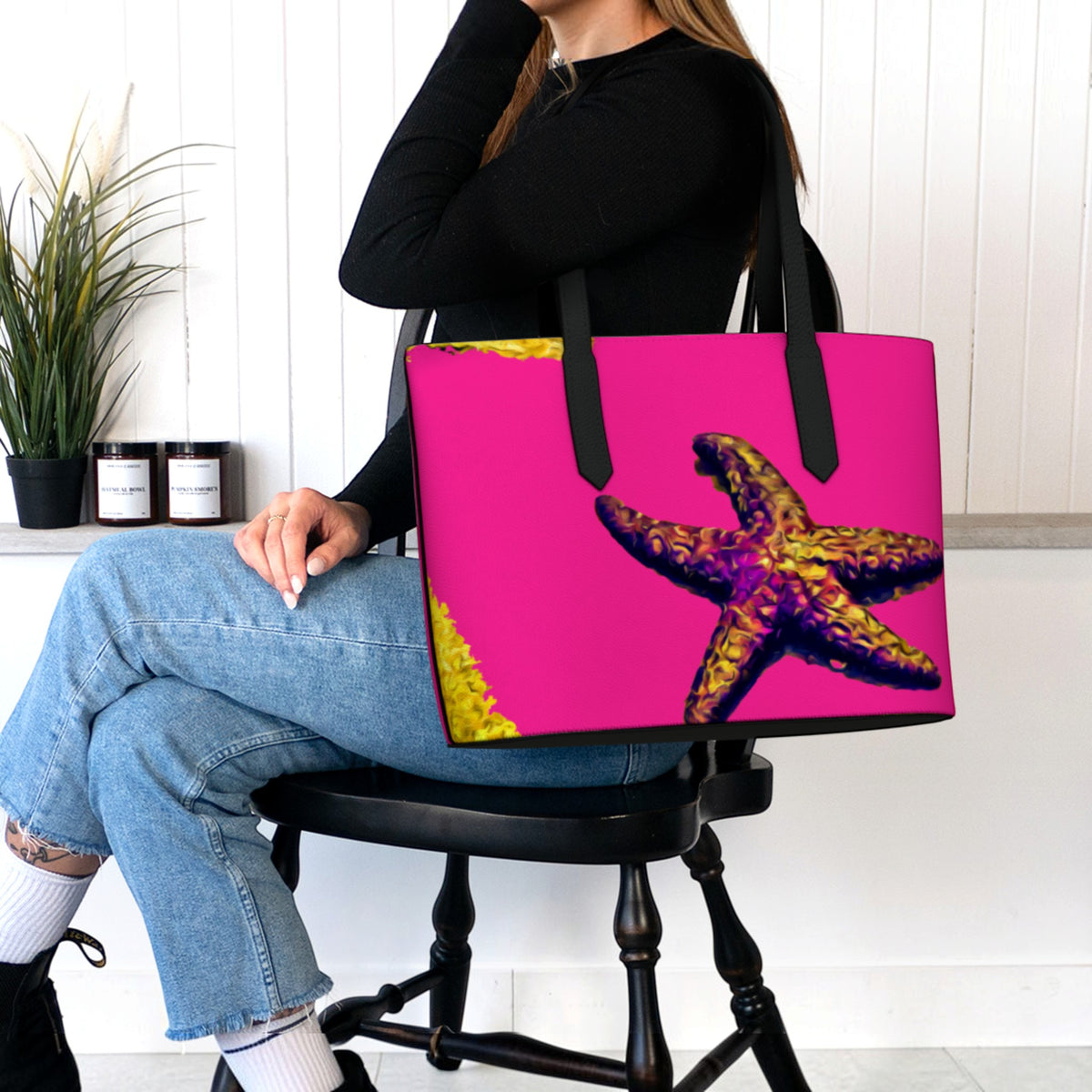 A woman sitting on a black wooden chair holds a large tote bag with a bright pink background and a large starfish print. The setting is a clean, modern room with white paneled walls, a potted plant, and candles.