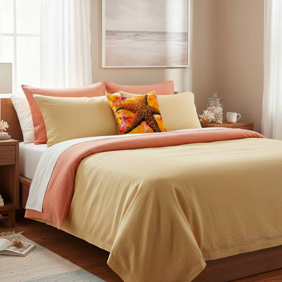 A neatly made bed in a softly lit bedroom, styled with a mustard yellow duvet cover, peach and cream pillows, and a colorful starfish accent pillow. The room includes wooden bedside tables, sheer curtains, and a framed beach-style print on the wall.