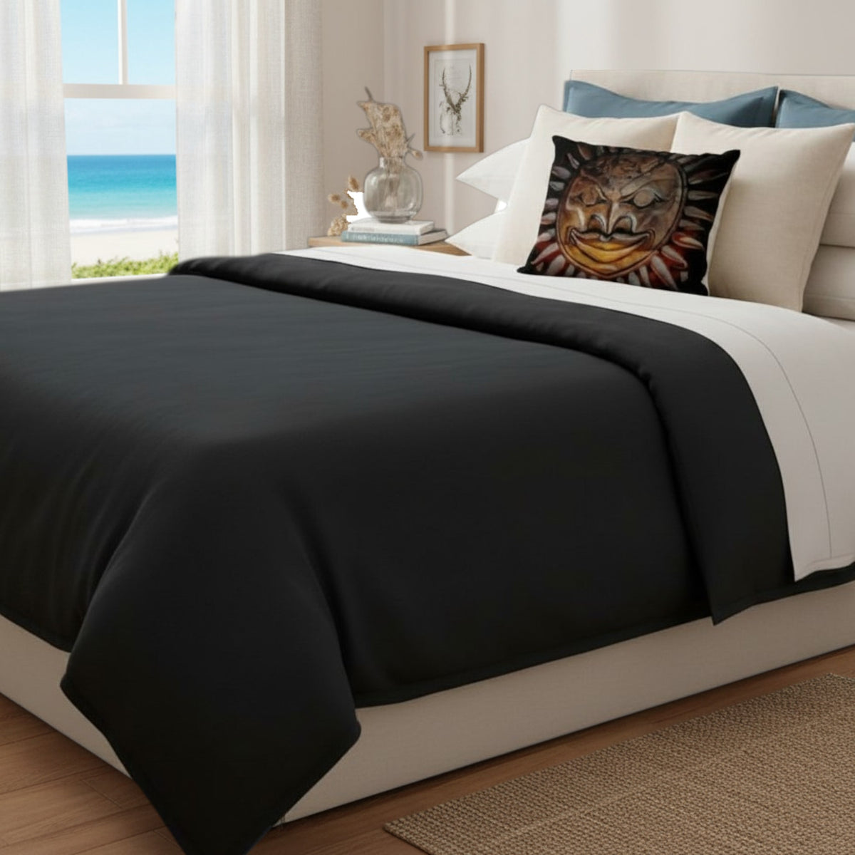 A bed dressed in black and white bedding, styled with a decorative sun-face accent pillow, set in a bright bedroom with a beach view through the window.