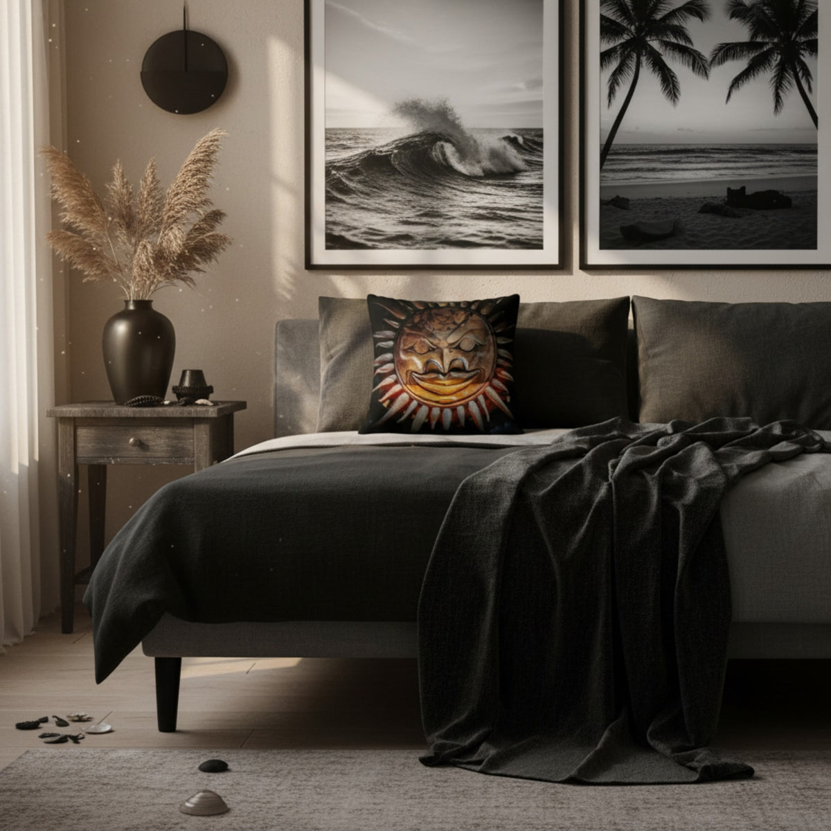 A styled bedroom scene with a bed dressed in dark charcoal bedding and a decorative throw blanket, featuring a square pillow with a colorful sun-and-moon face design. The room includes a side table with a vase of dried grass, framed black-and-white ocean wall art above the bed, and a gray rug on the floor.