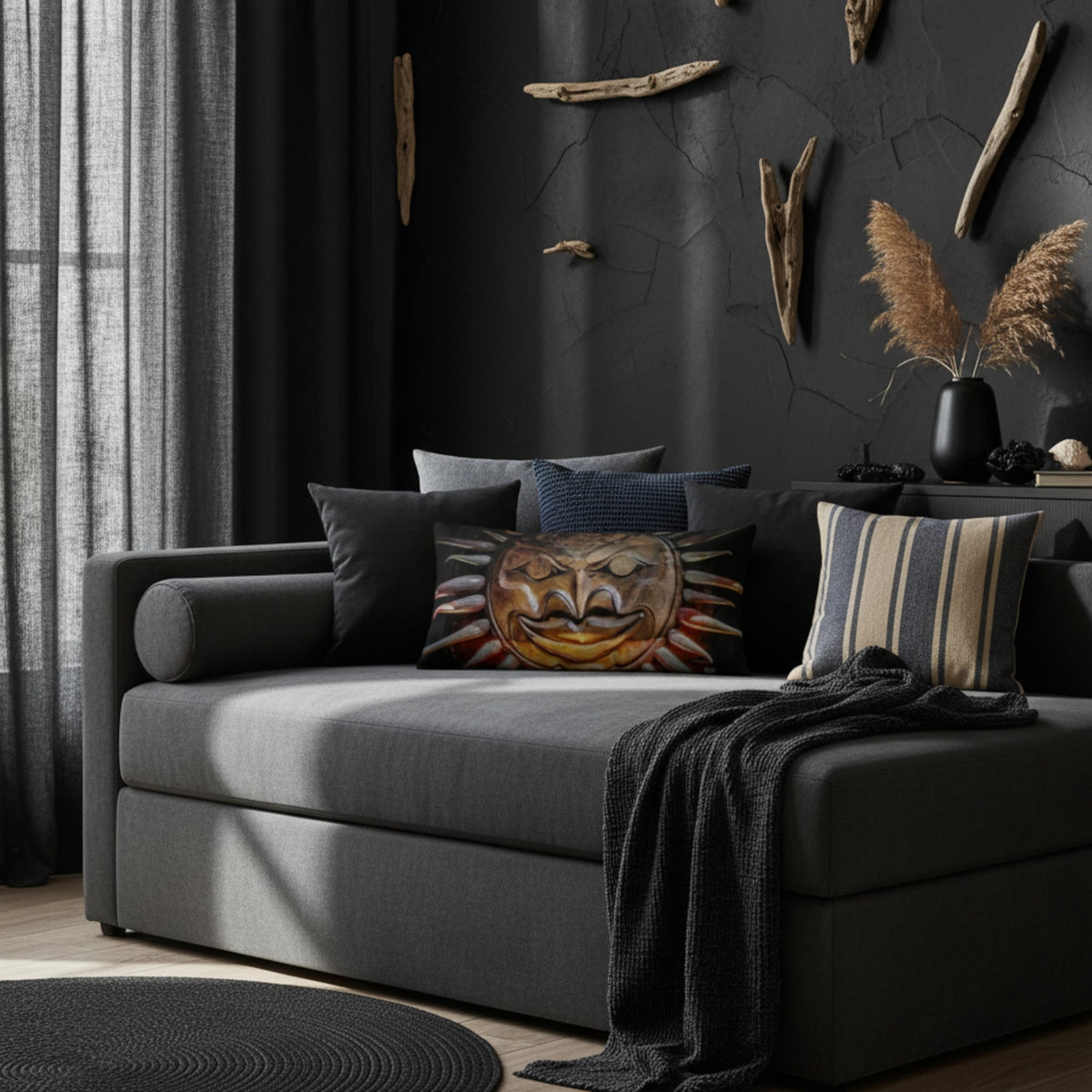 A dark gray upholstered daybed or chaise in a moody, rustic room setting. The bed is styled with several decorative throw pillows, including a central pillow featuring a carved wooden sun face design, and a dark charcoal knit throw blanket is draped over the front.