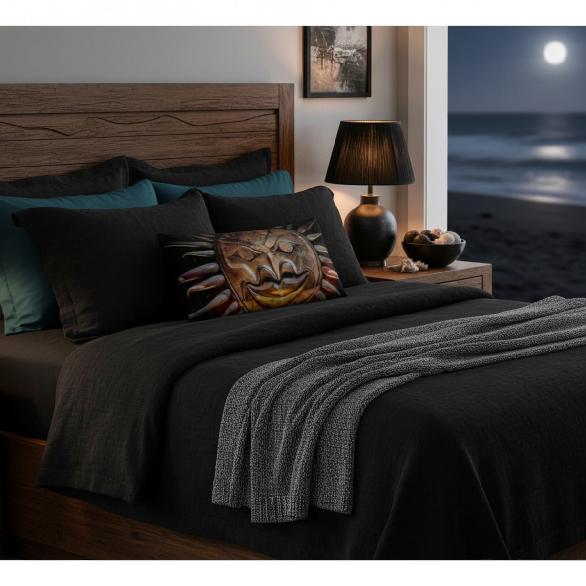 A styled bed in a bedroom setting with dark charcoal bedding, teal pillows, a decorative patterned accent pillow, a wooden headboard, bedside lamp, and a nightstand. A large window shows a beach scene at night under a full moon.