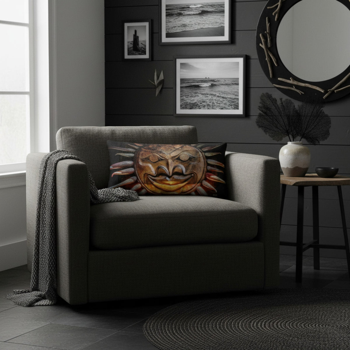 A gray upholstered armchair in a styled living room, featuring a square throw pillow with a colorful carved mask design. The room has a dark gray shiplap wall with framed black-and-white ocean photographs, a round mirror, and a small side table.