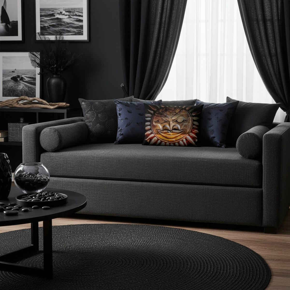 A dark gray upholstered loveseat in a moody black living room, styled with several throw pillows including a prominent sun-face patterned pillow, a round black coffee table, and dark curtains.