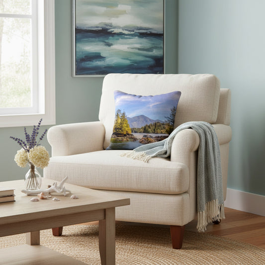 A cream upholstered armchair in a styled living room setting, featuring a square throw pillow with a colorful mountain landscape print and a draped gray throw blanket.