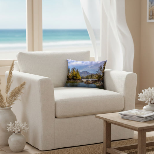 A square throw pillow featuring a scenic landscape print of a lake and mountains, styled on the seat of a cream-colored armchair in a bright, modern living room with an ocean view.
