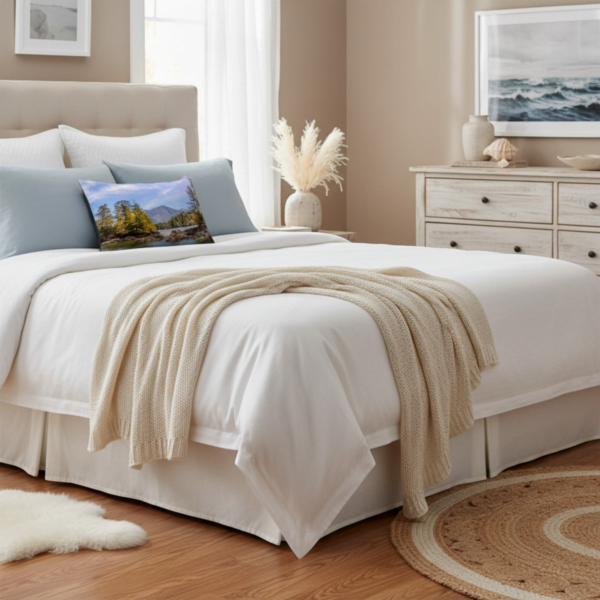 A neatly made bed in a softly styled bedroom, dressed in white bedding with a beige knit throw draped across the foot. Light blue pillows and a small landscape accent pillow are visible, with a dresser, framed wall art, and wood flooring in the background.
