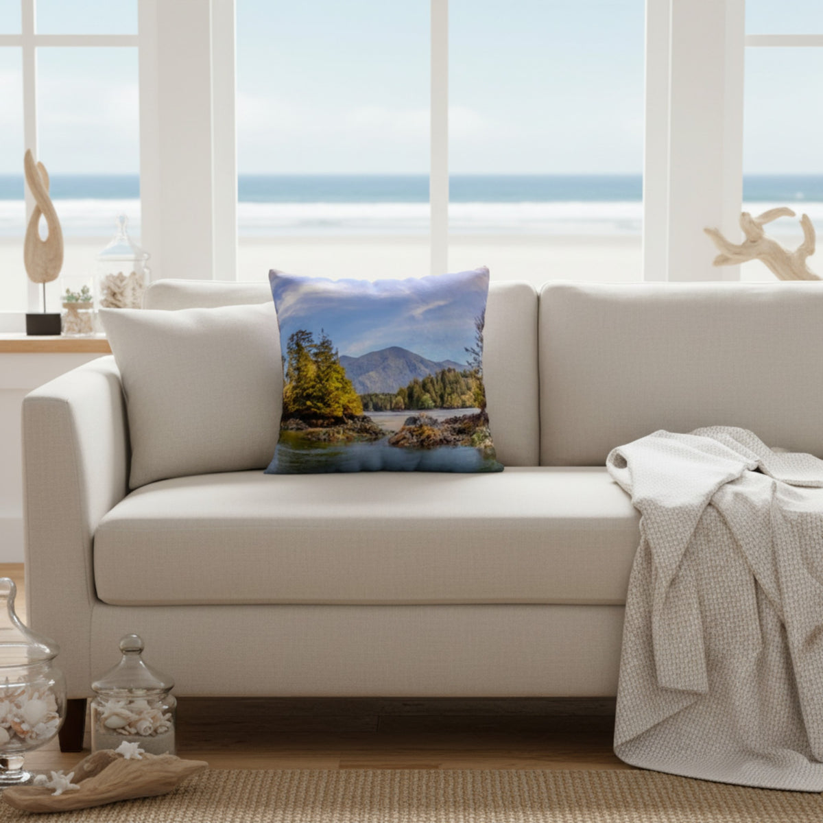A square throw pillow featuring a scenic landscape print of mountains, trees, and water, styled on a cream-colored sofa in a bright, modern living room with a beach view.