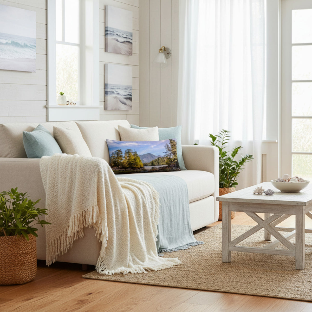 A bright, modern living room featuring a cream-colored sofa styled with a cream throw blanket, a light blue throw blanket, and a landscape print pillow. A white wooden coffee table sits on a natural jute rug, with potted green plants and white shiplap walls in the background.