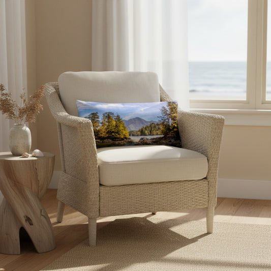 A square decorative throw pillow featuring a scenic landscape print of mountains, trees, and water, styled on a cream-colored woven armchair in a bright, modern room.