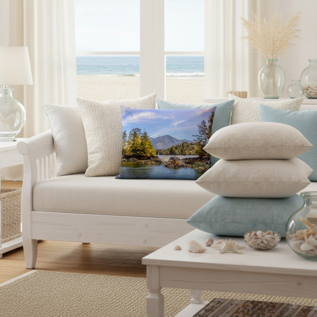 A square decorative throw pillow featuring a scenic landscape print of a lake, trees, and mountains, styled on a white sofa in a bright, coastal-themed living room.