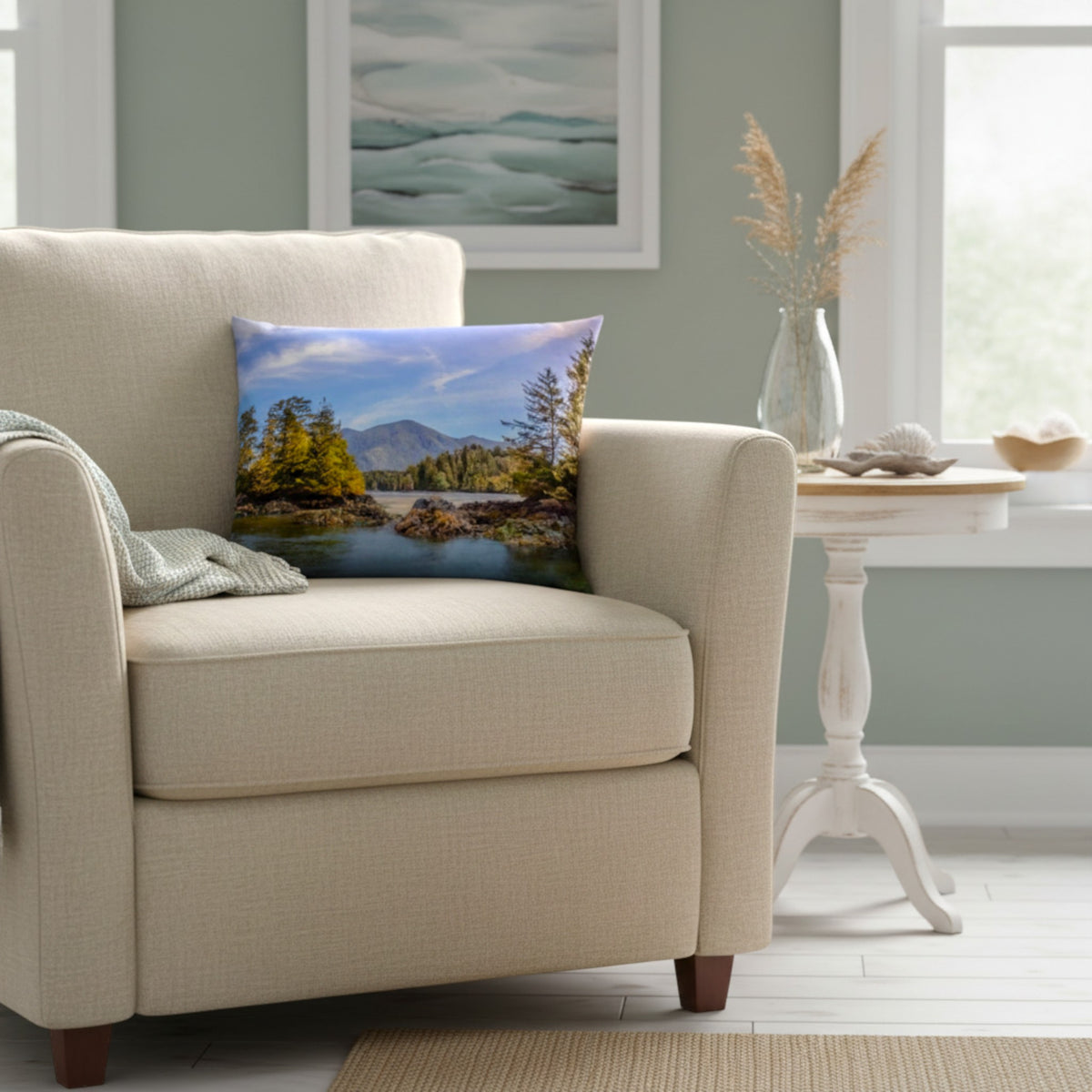 A square throw pillow featuring a scenic landscape print of a lake and mountains, styled on a beige armchair in a bright, modern living room.