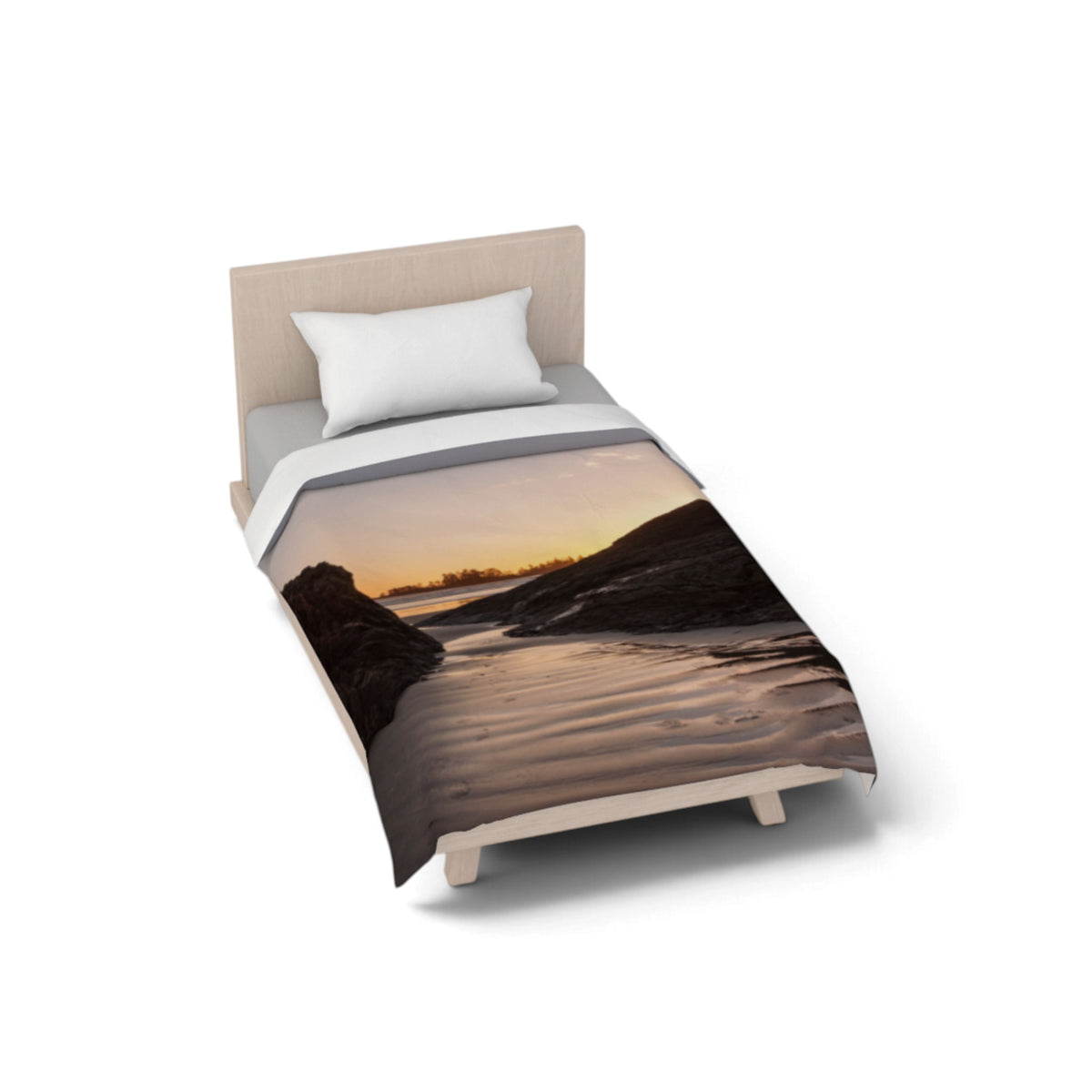 A single bed with a light wood frame and a white pillow, shown on a clean white background. The bed is dressed in a duvet cover featuring a scenic landscape print of a beach or river at sunset, with warm orange and yellow tones over dark rocky silhouettes.