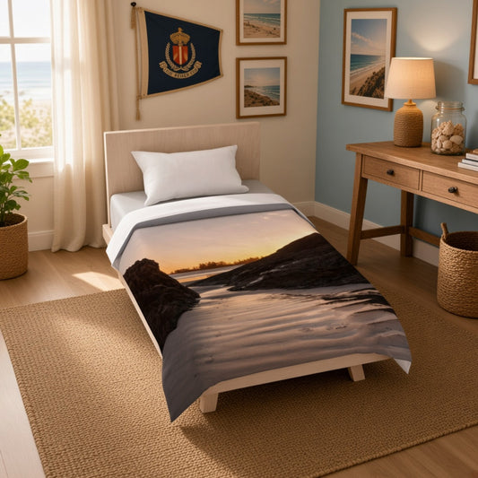 A single bed in a styled bedroom setting, shown with a duvet cover featuring a scenic landscape print of a beach shoreline at sunset. The room includes a wooden desk, lamp, framed beach photos, a flag, and a window with curtains.