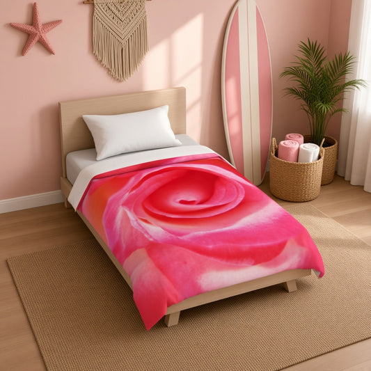 A single bed in a pink bedroom setting, featuring a vibrant pink and red rose print comforter. The room includes a wooden headboard, white pillow, a pink surfboard leaning against the wall, a potted plant, and a woven macrame wall hanging.
