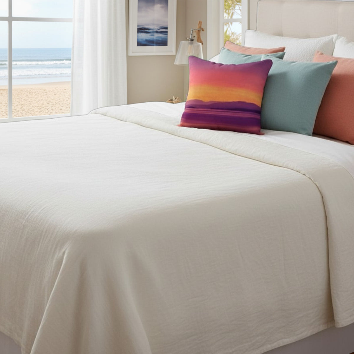 An off-white textured cotton sheet or duvet cover spread across a bed, styled with a colorful sunset-patterned accent pillow and other decorative pillows in a bright bedroom with a beach view through the window.