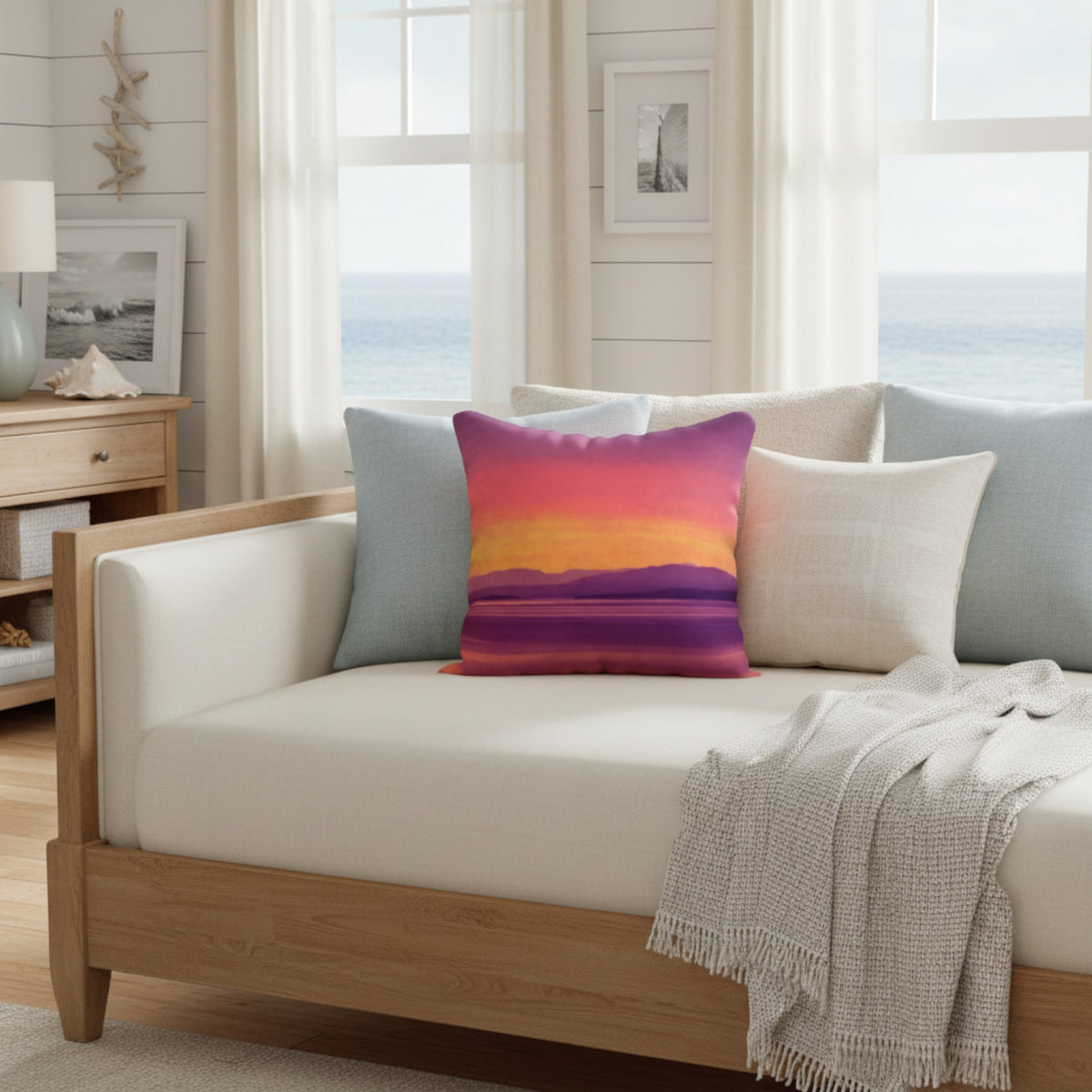 A square throw pillow with a colorful sunset landscape print in purple, pink, orange, and yellow, styled on a light wood daybed with white cushions in a bright, coastal-themed room.