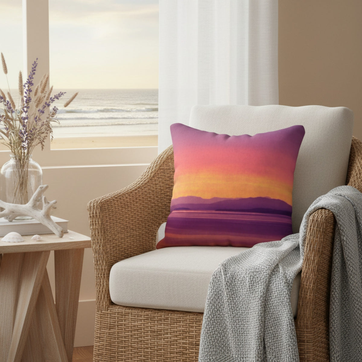 A square throw pillow with a colorful sunset landscape print in purple, pink, orange, and yellow, resting on a white cushion in a wicker armchair with a gray throw blanket, in a coastal living room setting.