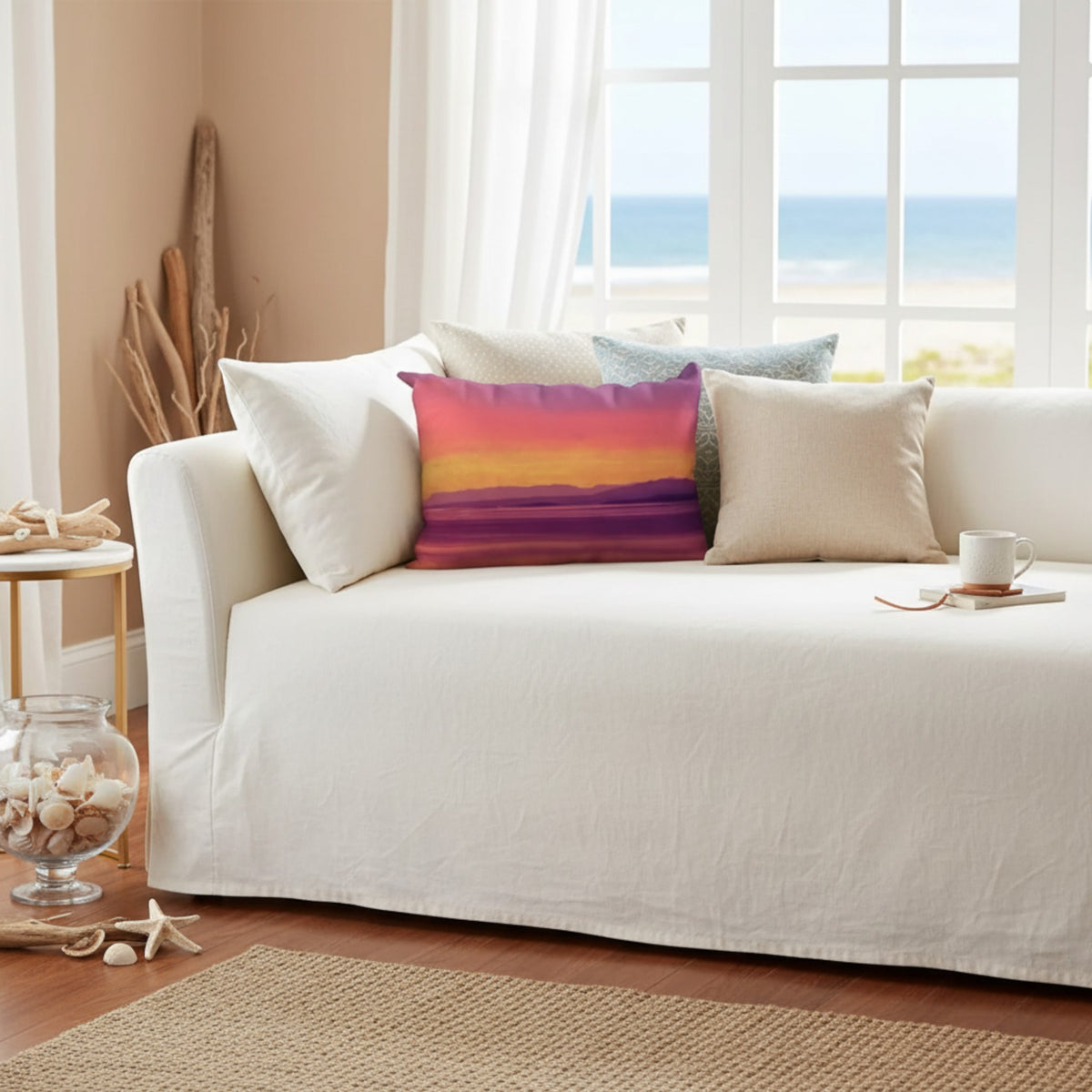 A white slipcovered daybed in a bright beach-themed living room, styled with several throw pillows including a prominent rectangular pillow featuring a sunset gradient in purple, orange, and yellow.