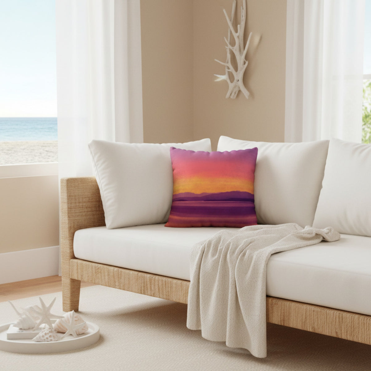 A square throw pillow featuring a colorful sunset landscape print in shades of pink, orange, yellow, and purple, styled on a white cushioned beach chair in a bright, modern living room.