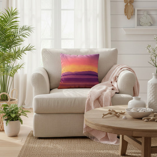 A square throw pillow with a colorful sunset landscape print in purple, pink, orange, and yellow, styled on a cream armchair in a bright, modern living room.