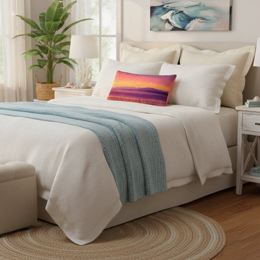 A neatly made bed in a bright bedroom setting, dressed in white bedding with a light blue textured throw blanket and a square accent pillow featuring a colorful sunset gradient.
