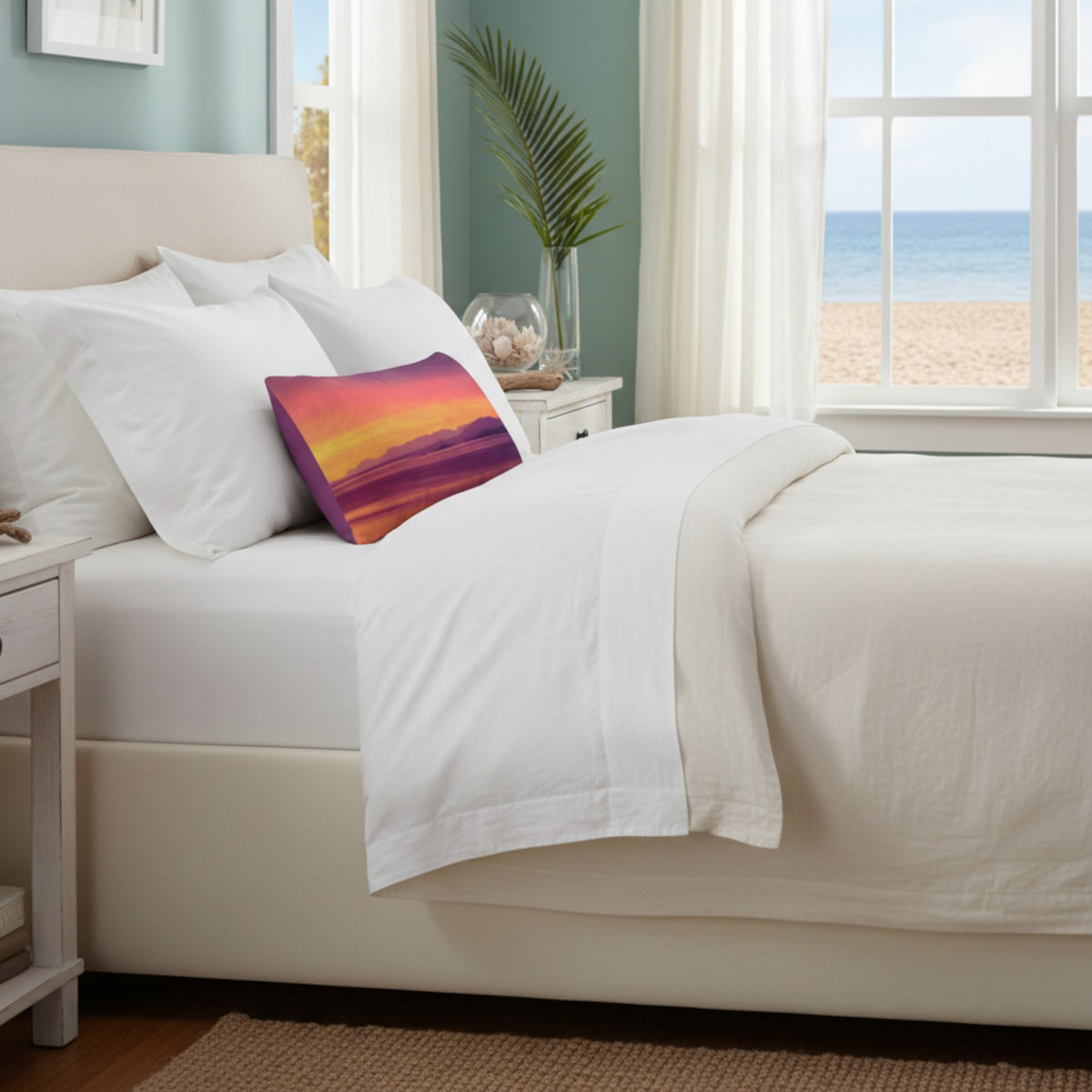 A neatly made bed with white bedding and a colorful rectangular accent pillow featuring a sunset gradient, set in a bright bedroom with a beach view through the window.