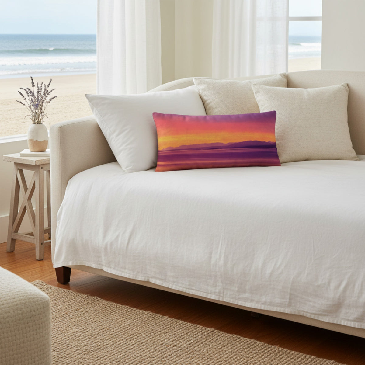 A rectangular lumbar pillow featuring a colorful sunset landscape print in purple, orange, and yellow, styled on a white bed in a bright beach-view bedroom.