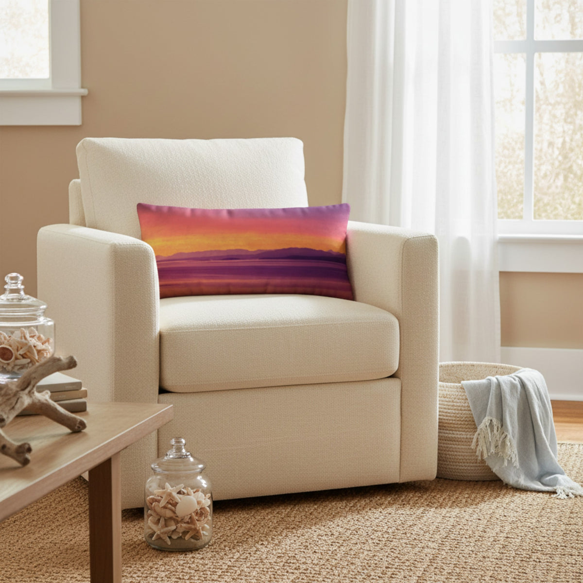 A cream upholstered armchair in a bright living room setting, styled with a rectangular throw pillow featuring a sunset landscape design in shades of pink, orange, yellow, and purple.