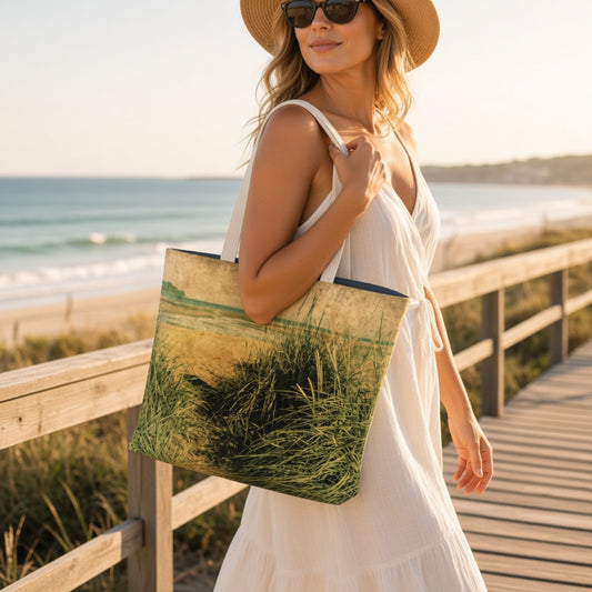 A woman in a white dress and straw hat carries a canvas tote bag with a beach grass landscape print, standing on a wooden boardwalk by the ocean.